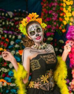 A lady with her face painted wearing a flower crown, black dress, and yellow lays.