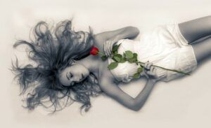 A black and white photo of a lady laying on the ground holding a rose. The rose is colored red.